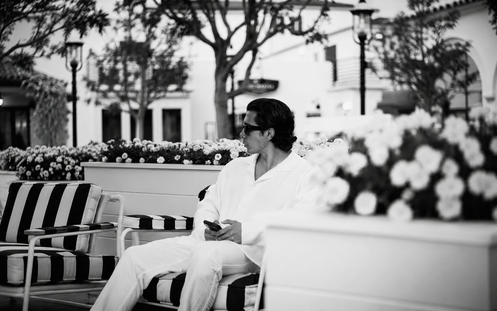 The founder seated on a striped terrace chair in white, looking off to the side, phone in hand.